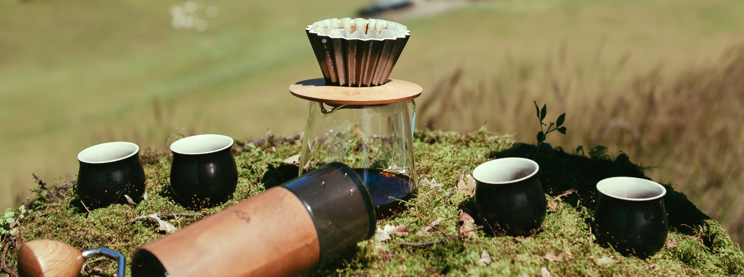 A pour over coffee maker and accessories in a lush green grass field