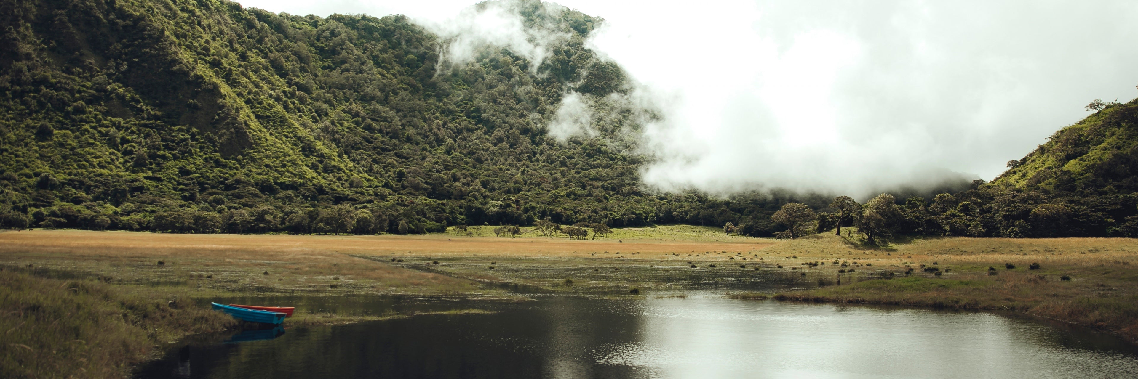 A small lake with two small boats set against a lush green mountain and rolling grey clouds. 